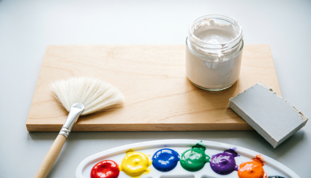 Wood painting supplies including a birch panel, gesso, sandpaper, and acrylic paints on a desk.